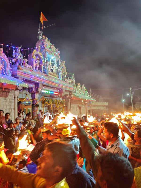 ಬಣ್ಣದ ದೀಪಗಳಿಂದ ಬೆಳಗುವ ದೇವಸ್ಥಾನದ ಗೋಪುರ ಮತ್ತು ಕೇಸರಿ ಧ್ವಜ; ಕೆಳಗೆ ಆರತಿ ಮೆರವಣಿಗೆ
