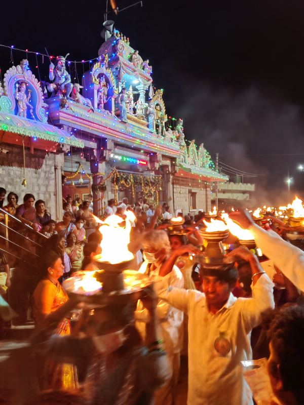 ದೀಪಾಲಂಕೃತ ದೇವಸ್ಥಾನದ ಮುಂಭಾಗ; ತಲೆಯ ಮೇಲೆ ಉರಿಯುವ ಆರತಿ ಹೊತ್ತ ಭಕ್ತರು