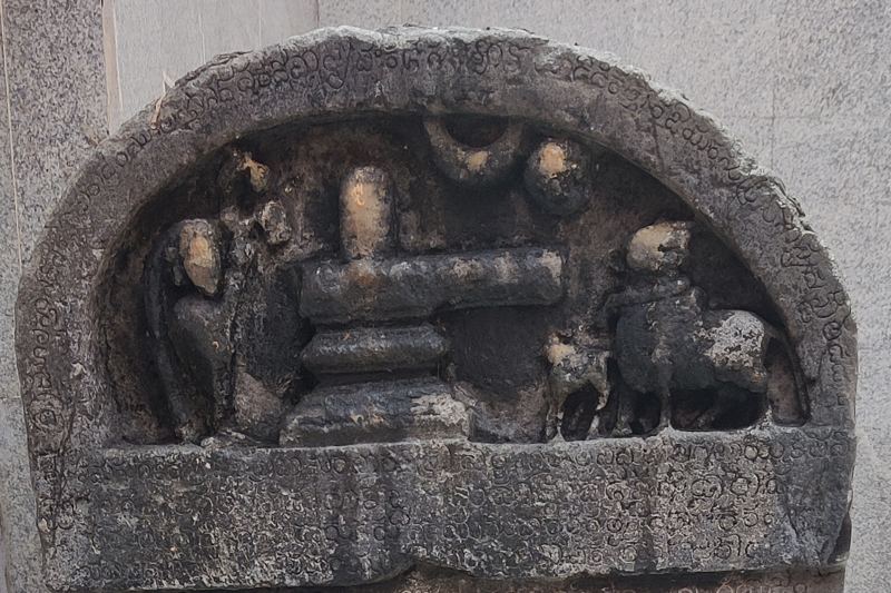 ಶಾಸನ ಕಲ್ಲಿನ ಮೇಲ್ಭಾಗದ ಶಿಲ್ಪ: ನಡುವೆ ಶಿವಲಿಂಗ, ಎಡಕ್ಕೆ ಕುಳಿತ ಭಕ್ತ, ಬಲಕ್ಕೆ ಕರುವಿನೊಂದಿಗೆ ಹಸು, ಮೇಲೆ ಚಂದ್ರ ಸೂರ್ಯರು; ಕಮಾನಿನ ಅಂಚಿನಲ್ಲಿ ಅಕ್ಷರಗಳು